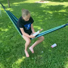 Person sitting in a hammock on grass, reading a book in a backyard setting.