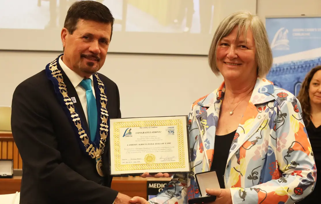 Photo of a woman with grey hair wearing colourful jacket accepting an award from a man with brown hair wearing a suit.
