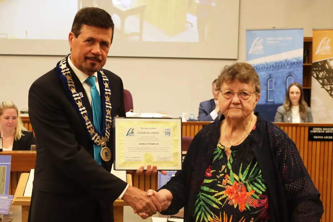 photo of a white woman in a floral shirt accepting an award from a man in a suit.