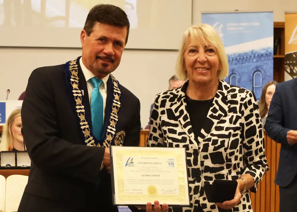 photo of a white woman in a suit,accepting an award from a man in a suit.