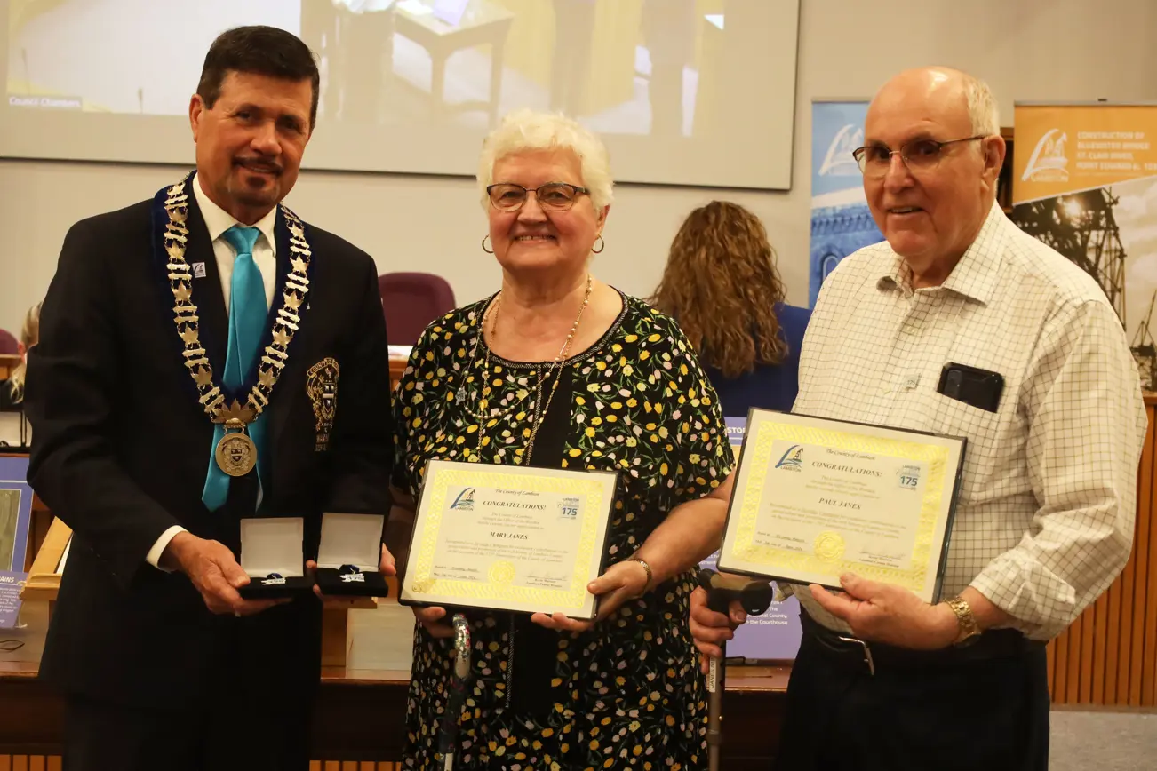 Photo of a woman with white hair and colourful shirt  and a man in a white plaid shirt accepting awards from a man in a dark suit.