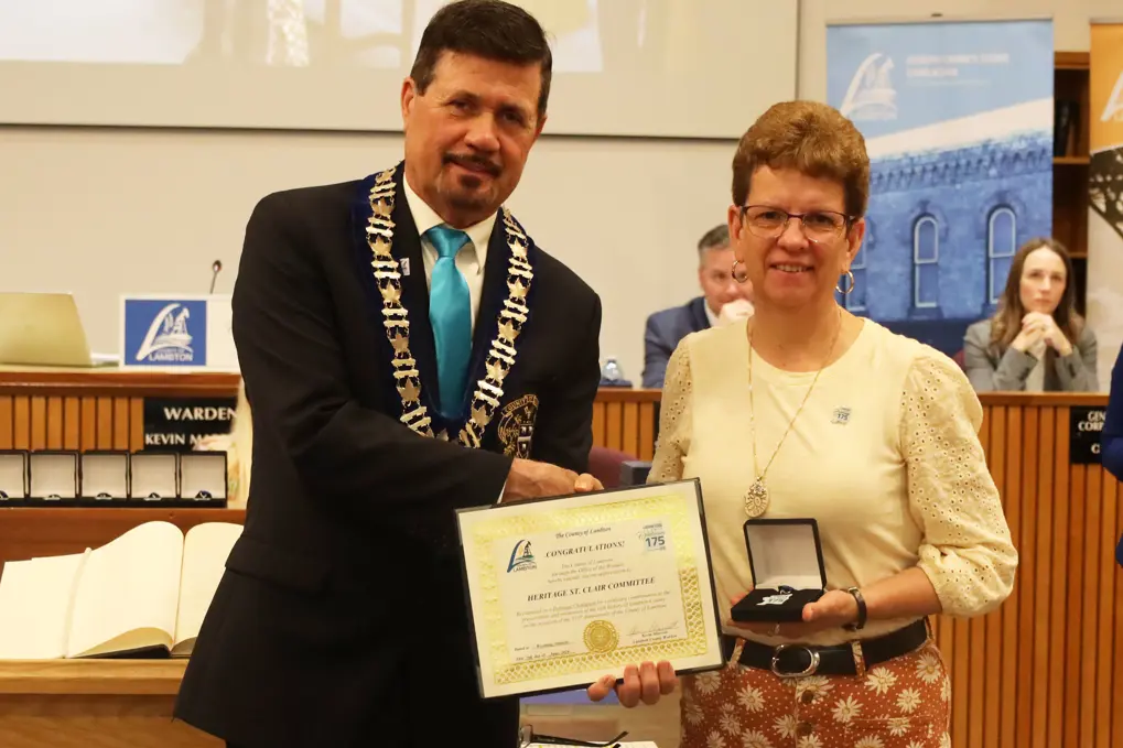 Photo of a woman in a white shirt and floral pants accepting an award from a man in a suit.